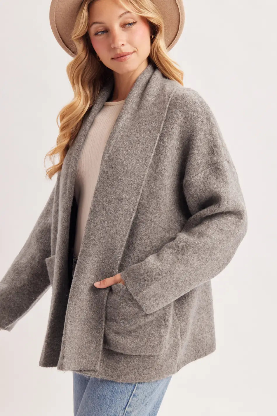 A woman wearing a light brown wide-brim hat, a gray open-front cardigan over a beige top, and blue jeans, photographed against a plain light gray studio background.