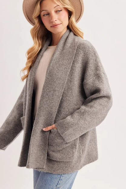 A woman wearing a light brown wide-brim hat, a gray open-front cardigan over a beige top, and blue jeans, photographed against a plain light gray studio background.