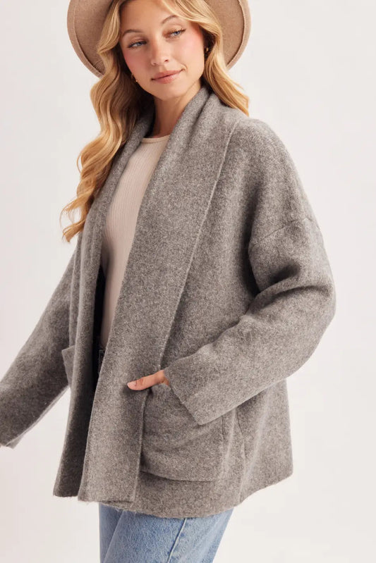 A woman wearing a light brown wide-brim hat, a gray open-front cardigan over a beige top, and blue jeans, photographed against a plain light gray studio background.