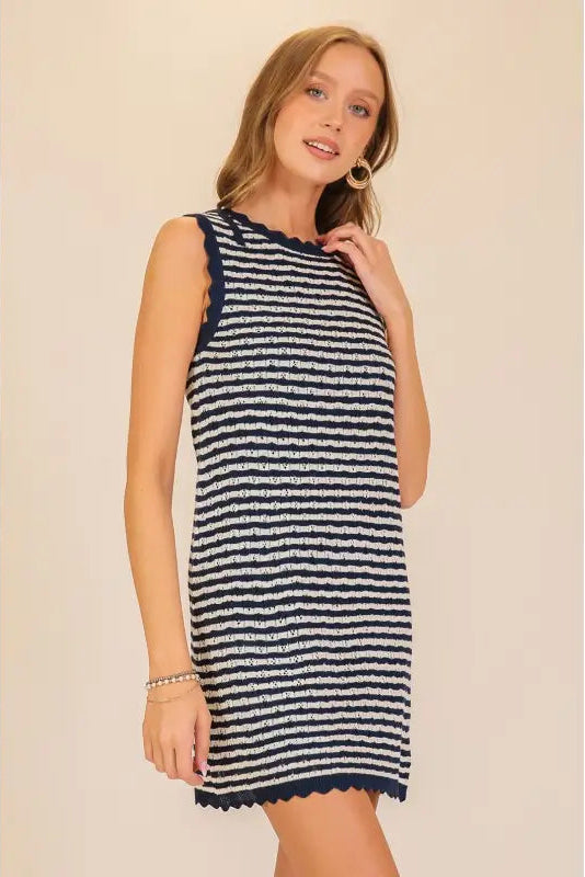 A woman modeling a sleeveless knit mini dress with horizontal navy and off-white stripes and a scalloped hem, set against a plain beige background.
