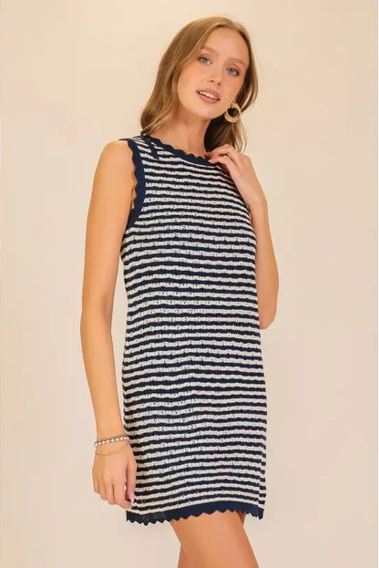 A woman modeling a sleeveless knit mini dress with horizontal navy and off-white stripes and a scalloped hem, set against a plain beige background.