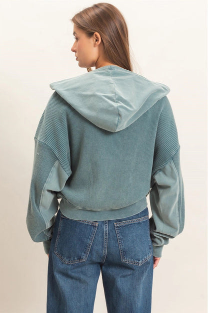 A woman shown from the back wearing a sage green corduroy cropped hoodie with textured panel details on the sleeves and hood, paired with blue jeans, against a plain off-white background.