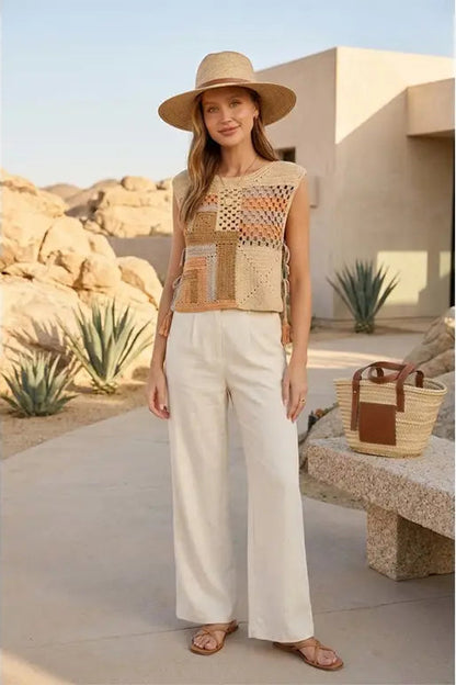 A woman stands outdoors in a desert-like setting wearing a sleeveless patchwork knit top with cream, tan, and orange tones, white wide-leg pants, a wide-brim straw hat, and flat sandals. A woven tote bag rests on a stone bench beside her.