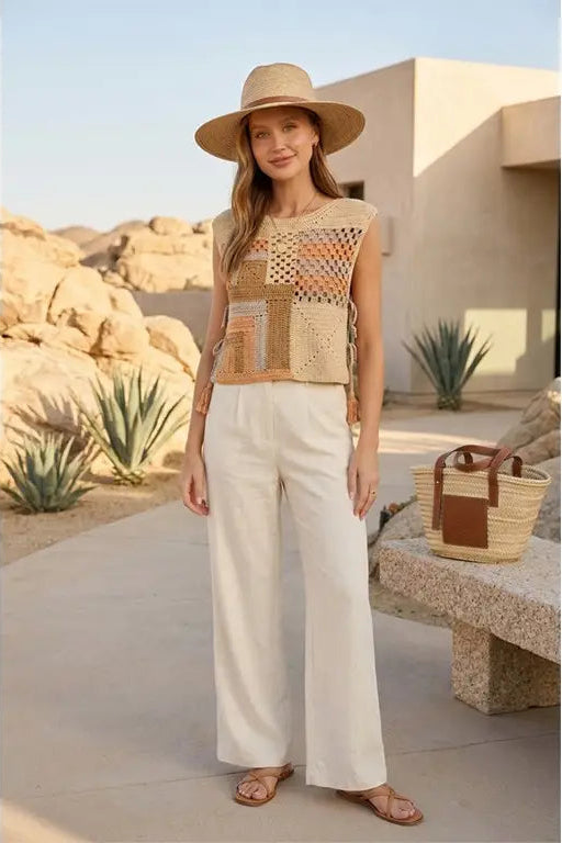 A woman stands outdoors in a desert-like setting wearing a sleeveless patchwork knit top with cream, tan, and orange tones, white wide-leg pants, a wide-brim straw hat, and flat sandals. A woven tote bag rests on a stone bench beside her.