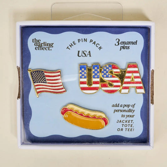 Set of three enamel pins featuring an American flag, 'USA' text, and a hot dog in a bun, displayed in a box by 'the darling effect'.