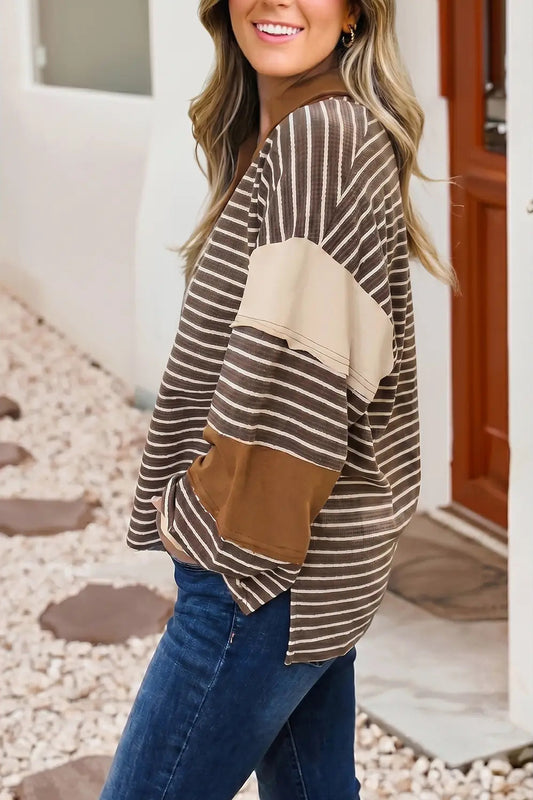 A woman wearing a brown and beige striped long-sleeve top with solid beige and brown color-block panels, paired with blue jeans, standing in front of a white wall and wooden door.