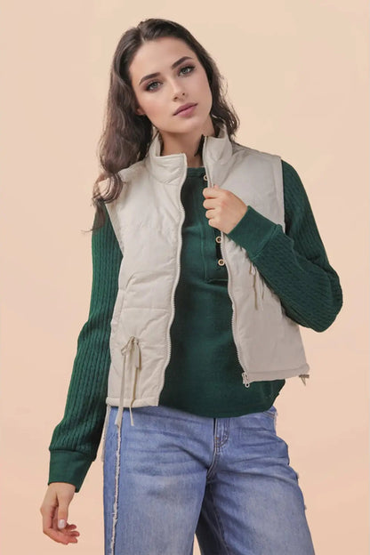 A woman modeling a sleeveless quilted puffer vest in light beige, worn over a dark green ribbed knit sweater and blue jeans, against a solid beige background.