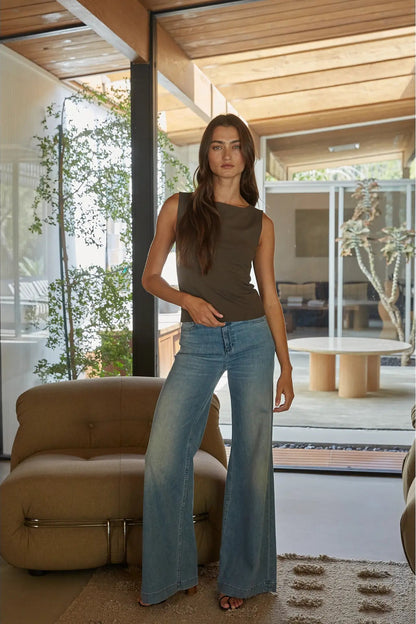A woman modeling a sleeveless olive-brown top and light-wash blue flare jeans in a modern indoor setting with large windows, wooden ceiling panels, and a tan armchair.