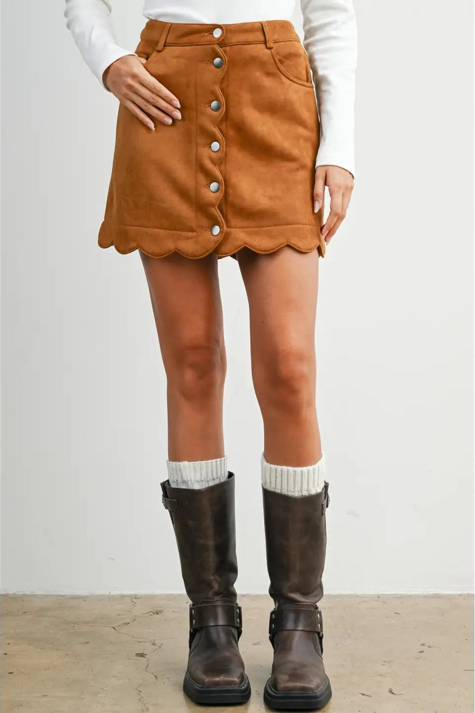 A model wearing a brown suede mini skirt with a scalloped hem and front button closure, styled with a white long-sleeve top, white socks, and dark brown leather boots.