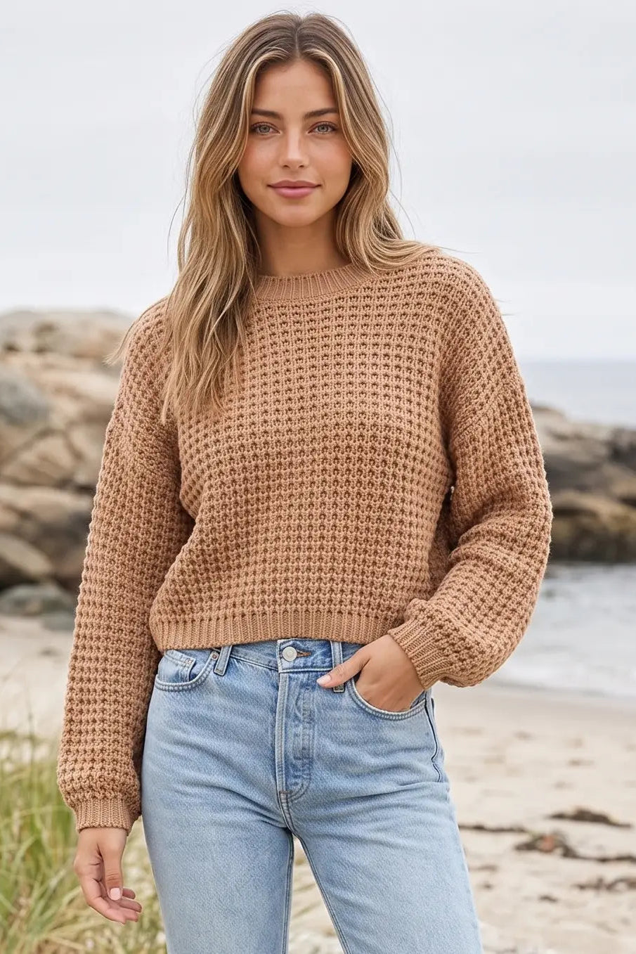 A woman standing on a beach wearing a textured, cropped waffle-knit sweater in a warm camel brown color, paired with light-wash blue jeans.