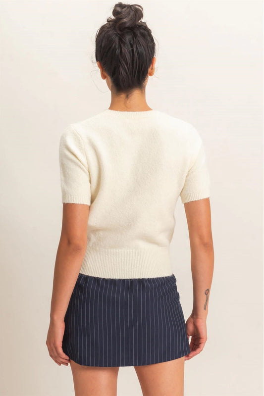 A woman shown from the back wearing a cream short-sleeve knit top and a navy blue pinstriped mini skirt, set against a plain light beige background.