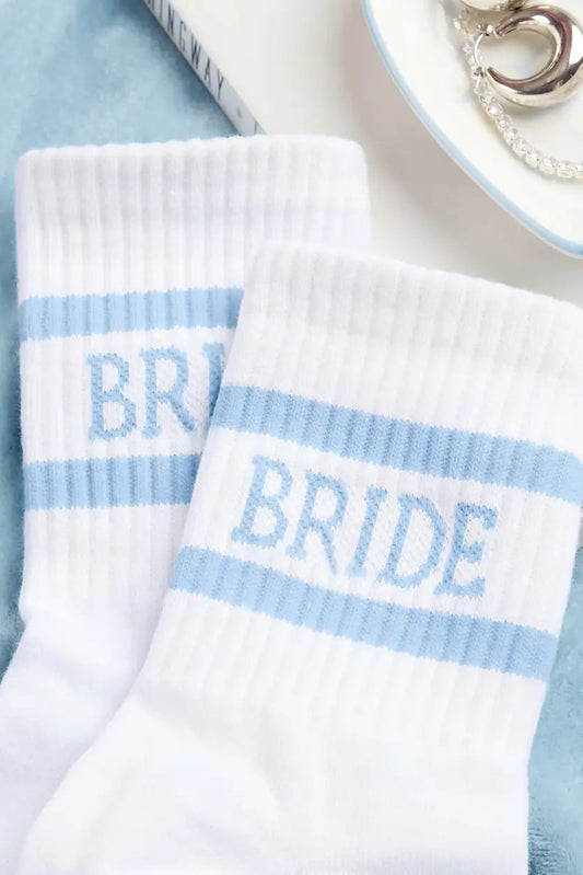 A pair of white crew socks with light blue horizontal stripes and the word 'BRIDE' knitted in light blue on the front sock, styled on a soft blue fabric background with a book and jewelry visible.