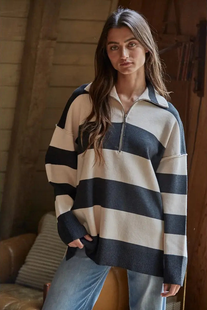A woman wearing an oversized knit half-zip sweater with wide horizontal navy and cream stripes, paired with light blue jeans, in a rustic indoor setting with wooden walls.