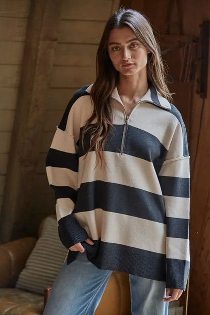 A woman wearing an oversized knit half-zip sweater with wide horizontal navy and cream stripes, paired with light blue jeans, in a rustic indoor setting with wooden walls.