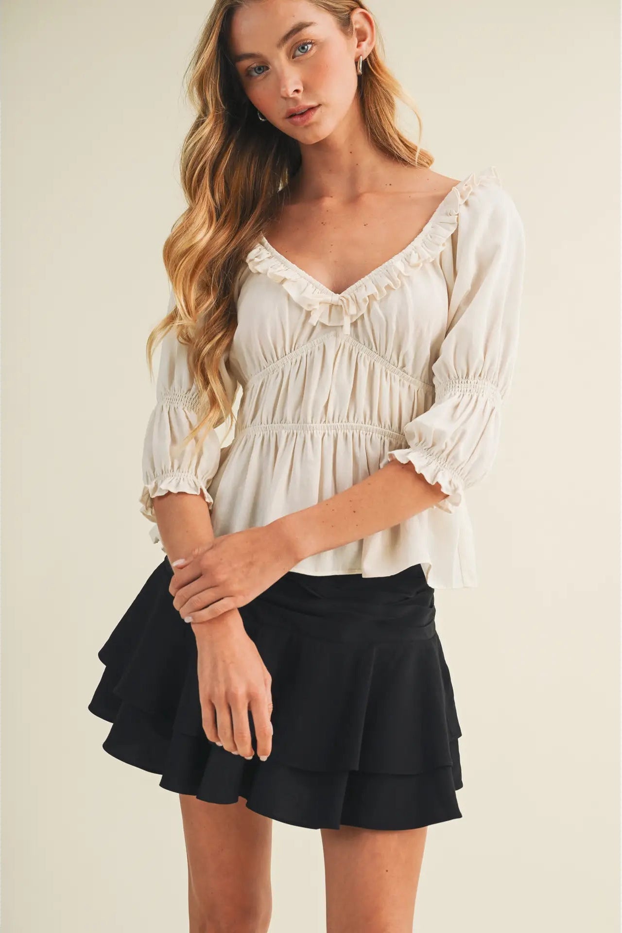 A woman modeling a cream-colored ruffled blouse with three-quarter sleeves and a black tiered ruffled mini skirt, set against a plain beige studio background.