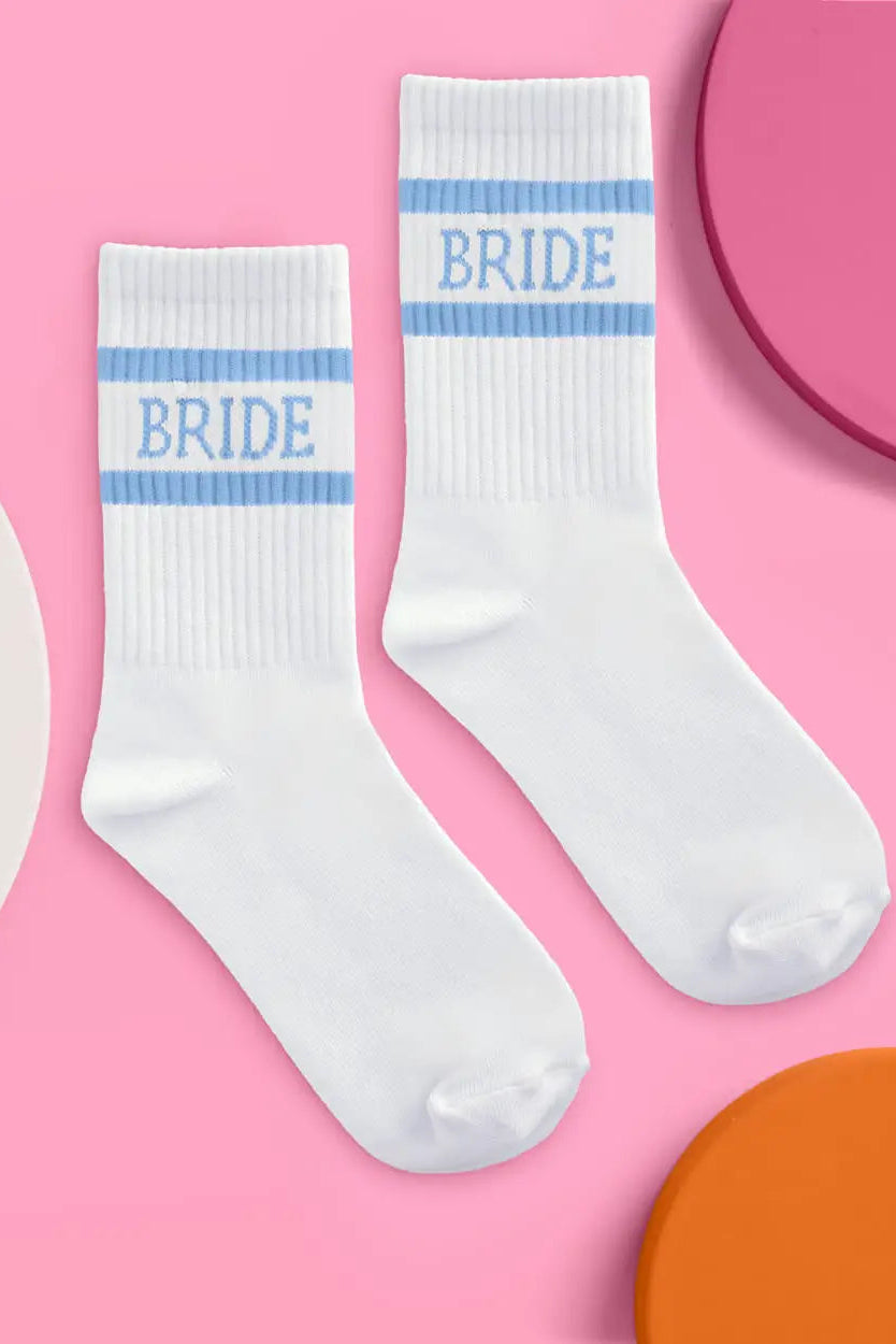 A pair of white crew socks with light blue stripes and the word 'BRIDE' printed in blue on the cuffs, arranged on a pink background with partial colorful circular props.