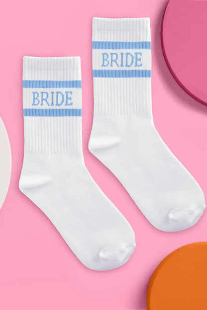 A pair of white crew socks with light blue stripes and the word 'BRIDE' printed in blue on the cuffs, arranged on a pink background with partial colorful circular props.