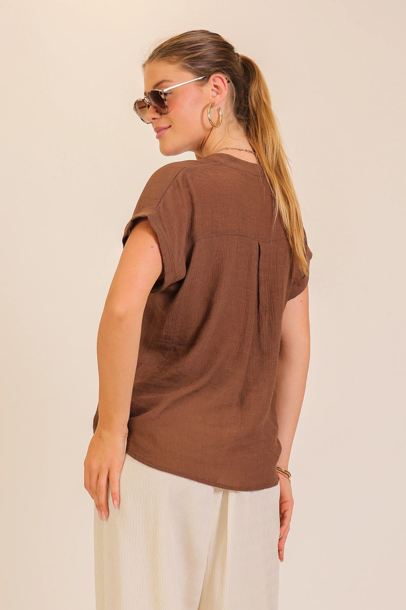 A woman wearing a brown short-sleeve top and white pants, photographed from the back against a plain light beige studio background.