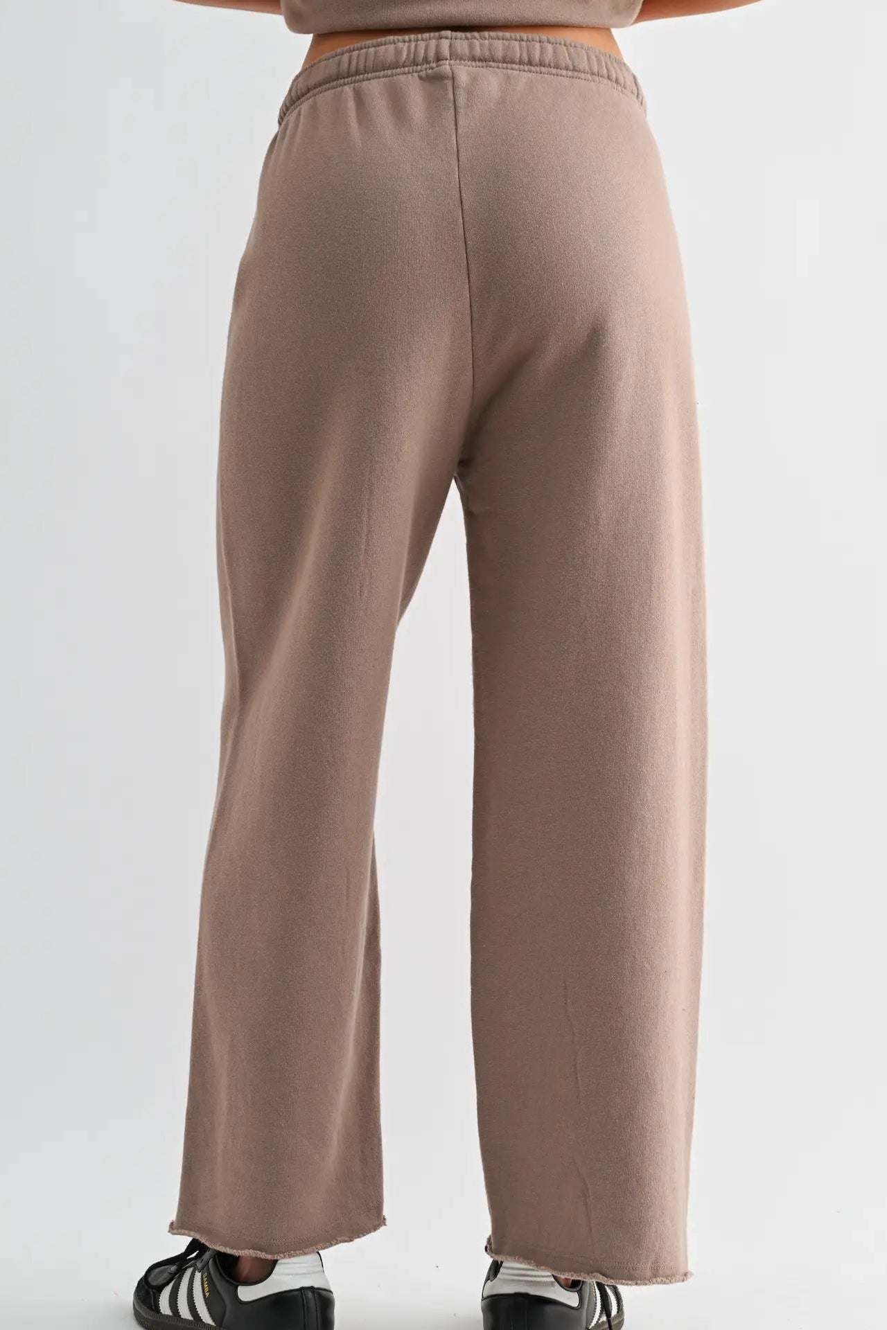 A person shown from the back wearing loose, wide-leg sweatpants in a muted taupe-brown color, paired with black and white sneakers against a plain light gray studio background.