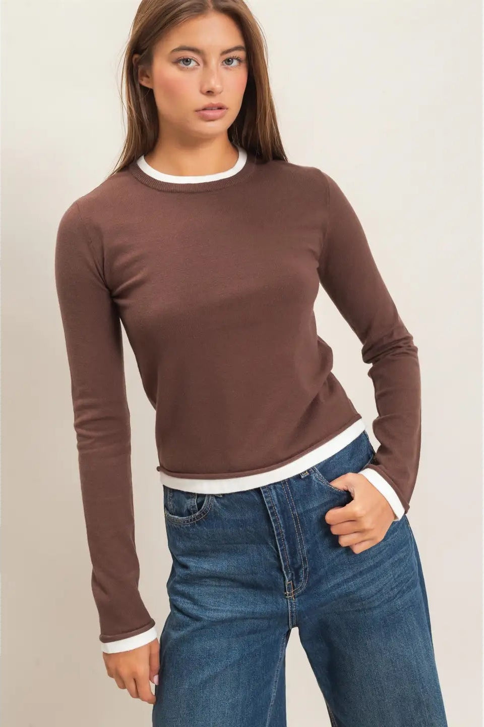 A woman modeling a fitted long-sleeve crewneck top in brown with white ribbed trim at the collar, cuffs, and hem, paired with blue jeans against a plain light background.