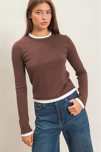 A woman modeling a fitted long-sleeve crewneck top in brown with white ribbed trim at the collar, cuffs, and hem, paired with blue jeans against a plain light background.