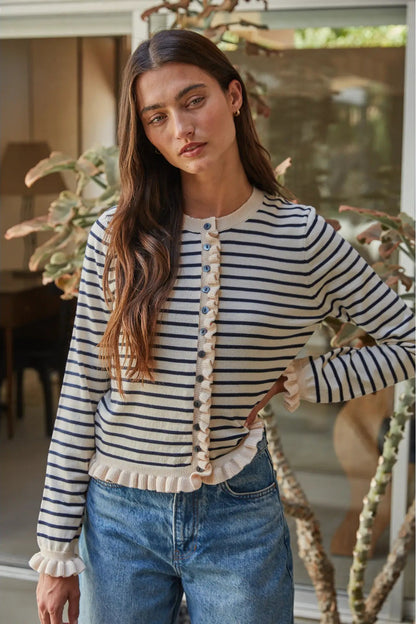 A woman wearing a cream and navy blue horizontally striped cardigan with a decorative button placket and ruffled trim, paired with blue jeans.