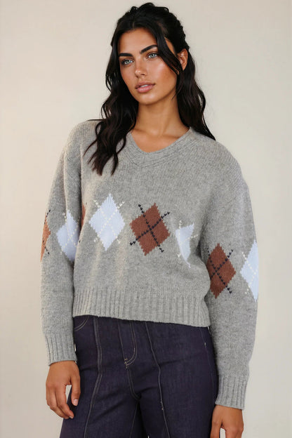 A woman wearing a light gray knit sweater with an argyle pattern in white, brown, and dark brown, paired with dark blue jeans, against a plain beige background.