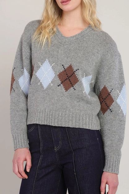 A woman wearing a gray knit sweater with an argyle diamond pattern in brown, light blue, and white, paired with dark blue jeans.