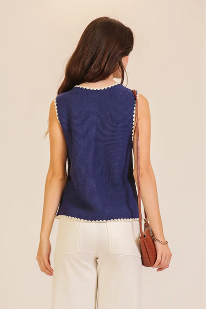 A woman shown from the back wearing a sleeveless navy blue knit top with white scalloped trim, paired with white pants and carrying a small brown shoulder bag, against a plain beige background.