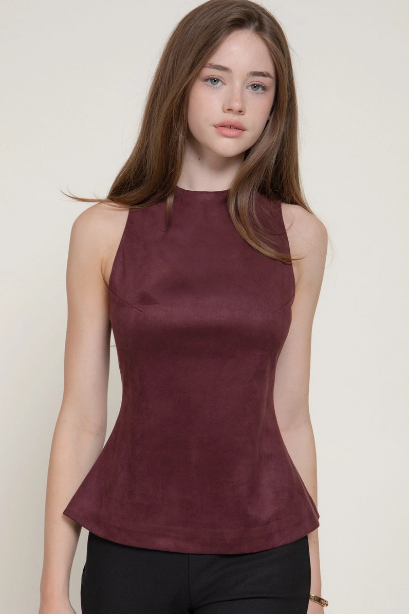 A woman wearing a sleeveless, high-neck peplum top in a deep burgundy color, paired with black pants, against a plain off-white background.