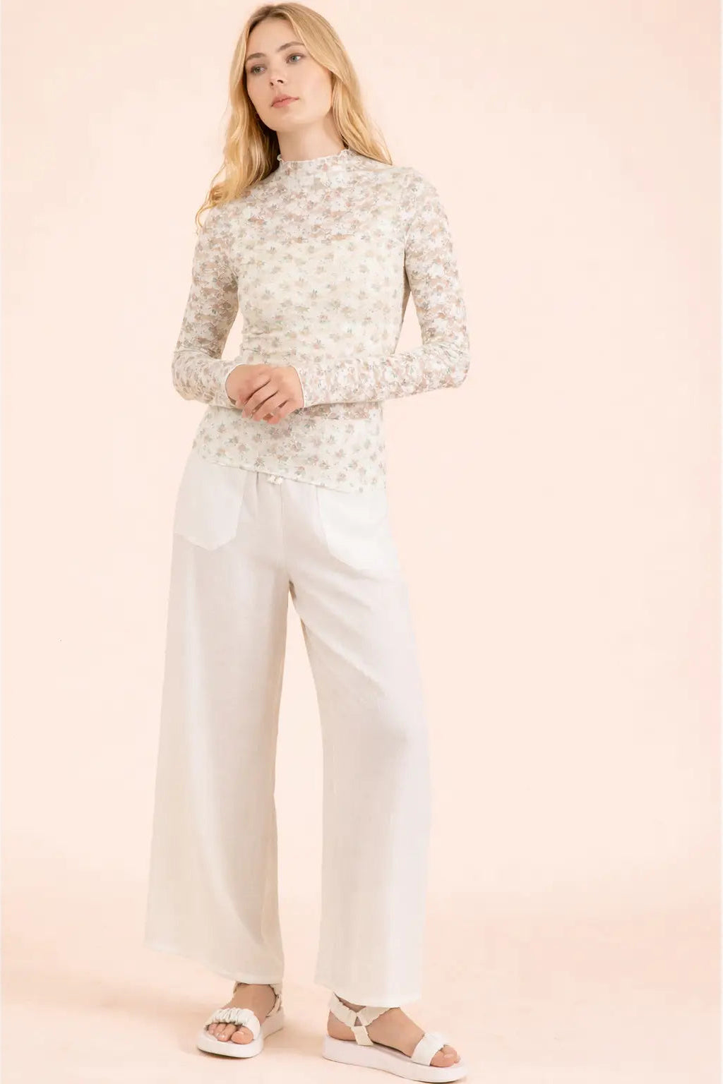 A woman modeling a long-sleeve mock-neck knit top with a delicate beige and white floral pattern, paired with white wide-leg trousers and white platform sandals, set against a solid pale pink background.