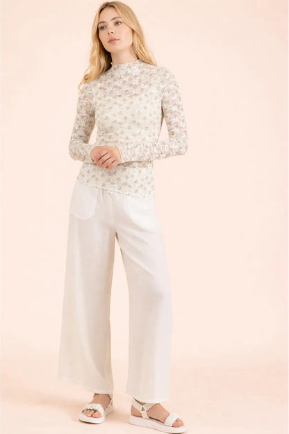 A woman modeling a long-sleeve mock-neck knit top with a delicate beige and white floral pattern, paired with white wide-leg trousers and white platform sandals, set against a solid pale pink background.