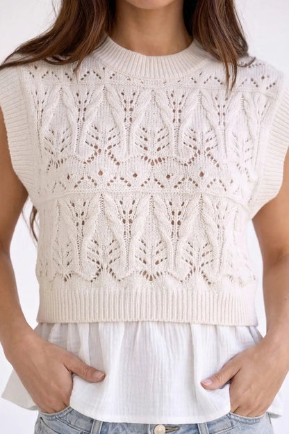 A woman wearing a sleeveless cream knit sweater vest with an intricate leafy cable-knit pattern over a white ruffled blouse and light blue jeans.