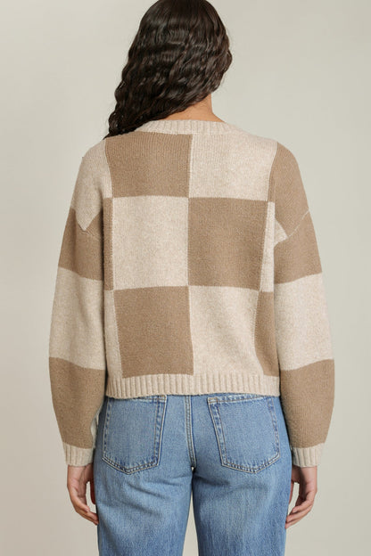 A woman viewed from the back wearing a beige and cream checkered knit sweater and blue jeans against a light gray studio background.
