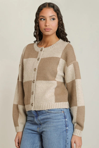A woman wearing a beige and cream checkered knit cardigan sweater with brown buttons, paired with blue jeans, against a solid light beige background.