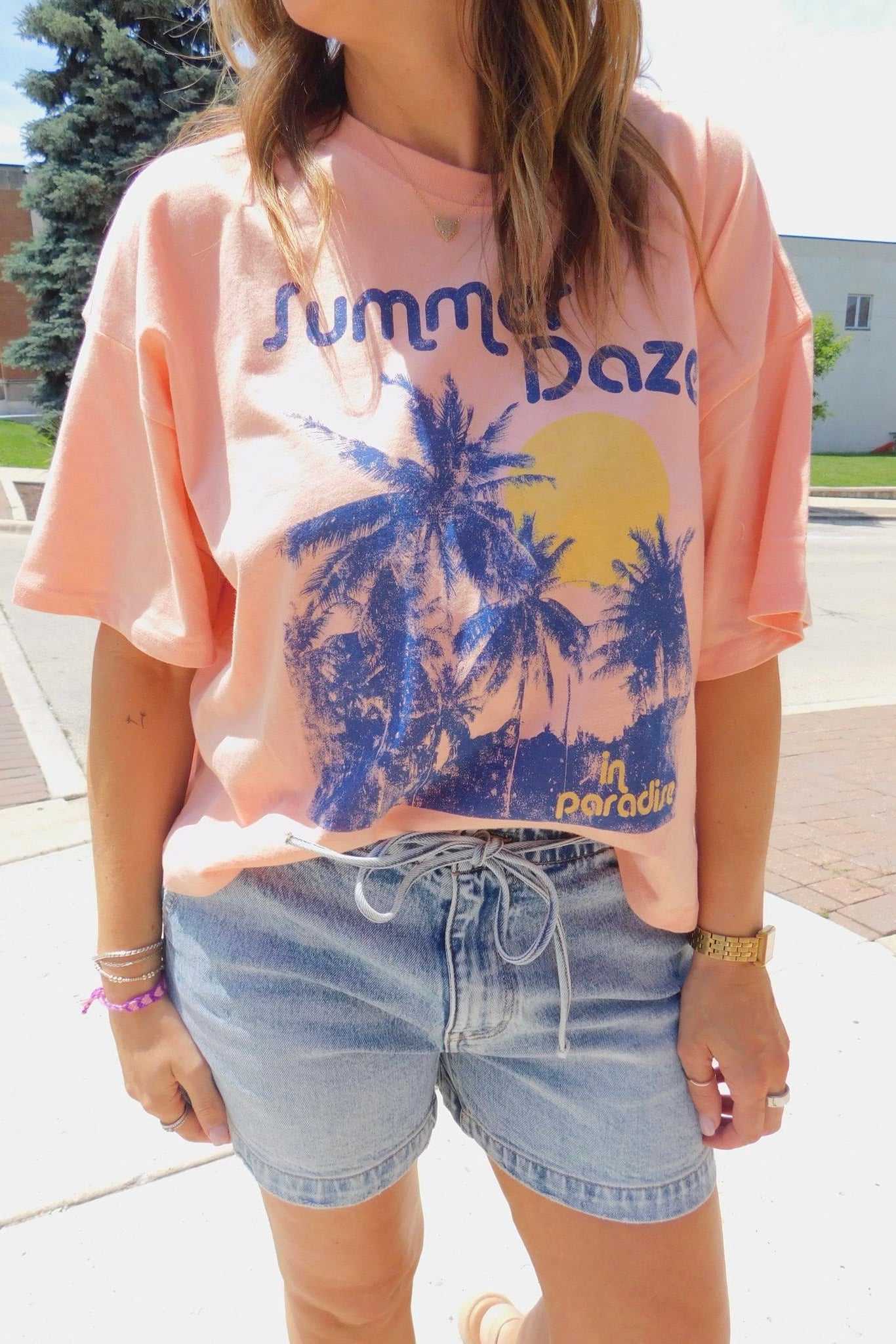 Person wearing a pink t-shirt with palm tree design and text, standing outdoors.