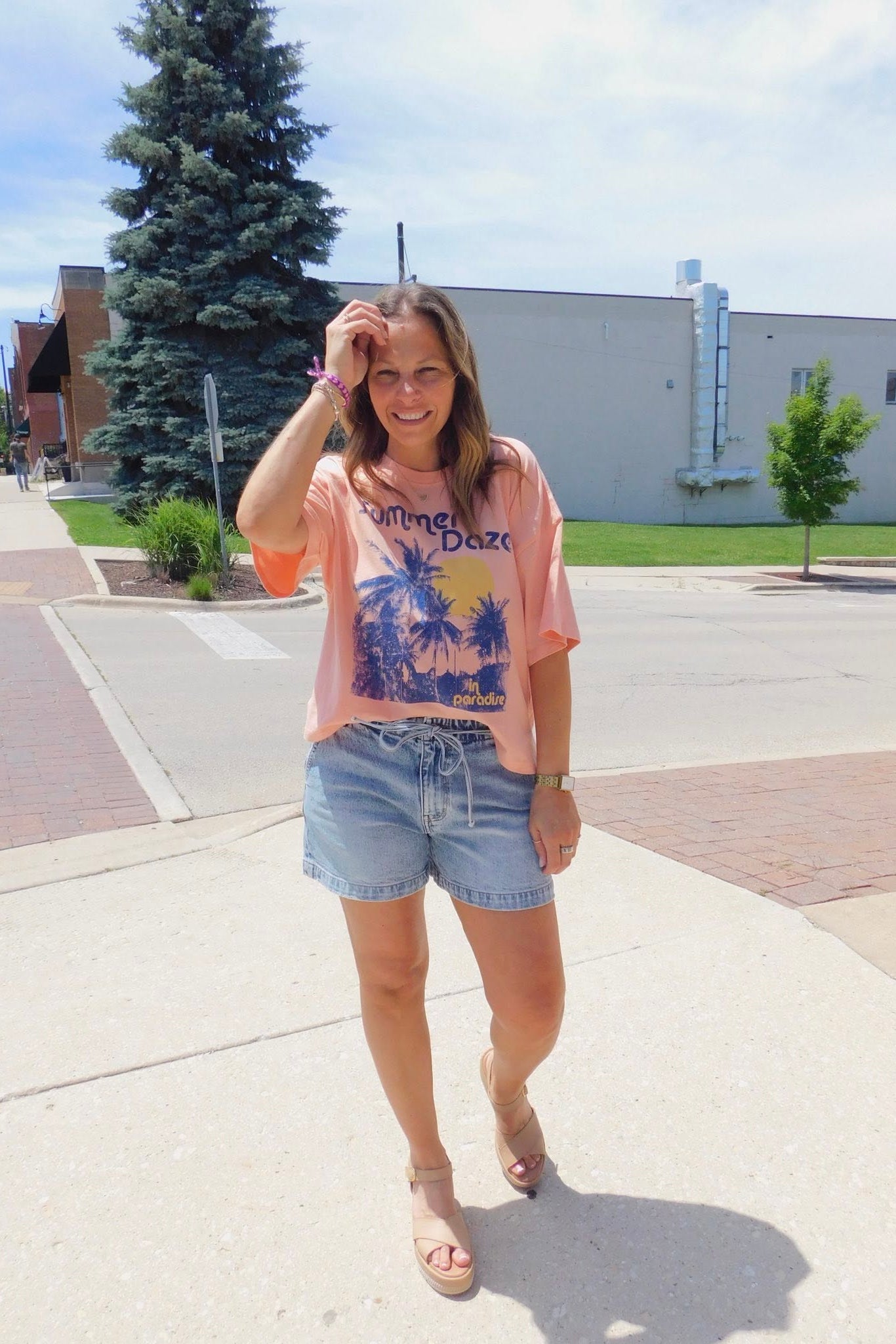 Woman in a pink graphic t-shirt and denim shorts standing on a sidewalk with trees and buildings in the background.