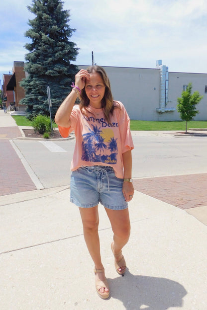 Woman in a pink graphic t-shirt and denim shorts standing on a sidewalk with trees and buildings in the background.