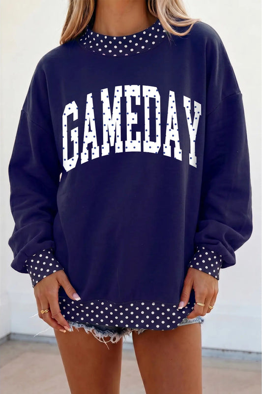 A woman wearing a navy blue crewneck sweatshirt with 'GAMEDAY' printed in white arched letters across the chest. The sweatshirt features white polka-dot trim around the neckline, cuffs, and hem, and is styled with frayed denim shorts.