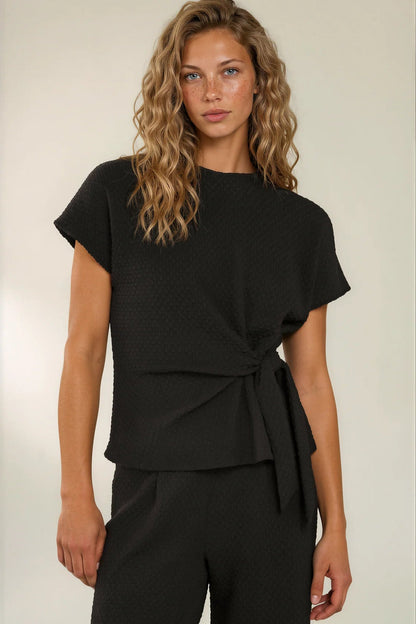 A woman modeling a black short-sleeve wrap top with a textured, waffle-like fabric, paired with matching black trousers, against a plain light beige studio background.