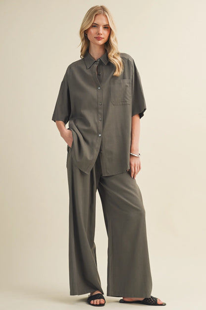 A woman modeling a matching two-piece outfit consisting of an oversized short-sleeve button-up shirt and wide-leg pants in a muted olive green color, set against a plain beige studio background.