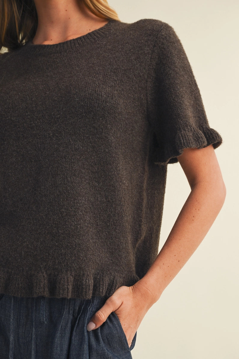 A close-up of a person wearing a dark brown short-sleeve knit sweater with ruffled cuffs and hem, paired with a blue skirt, against a plain light beige background.