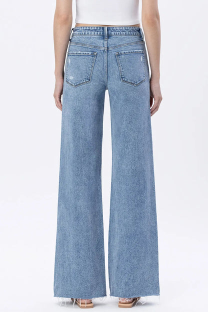 A model shown from the back wearing light blue wide-leg denim jeans with a frayed hem, paired with a white top and beige sandals, against a plain white studio background.