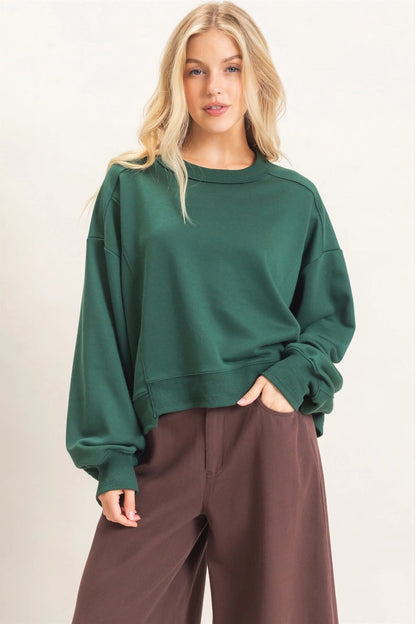 A blonde model wearing a dark green crewneck sweatshirt and brown wide-leg pants, standing against a plain off-white studio background.