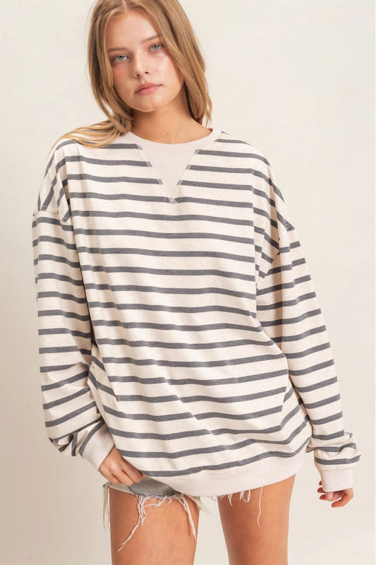 A woman wearing an oversized long-sleeve crewneck sweatshirt with horizontal gray stripes on a cream base, paired with frayed denim shorts, against a plain beige studio background.