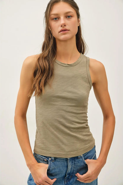 A woman modeling a sleeveless crew-neck tank top in a muted sage green color, paired with blue jeans, against a plain light gray studio background.