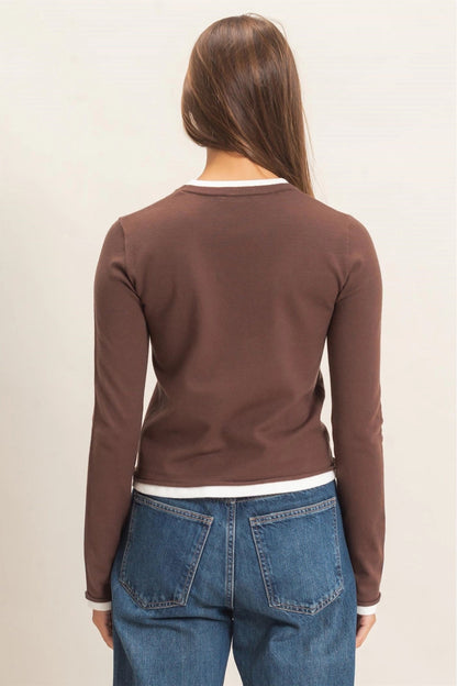 A woman viewed from the back wearing a fitted brown long-sleeve top with a white crew neck and blue jeans, against a plain light beige studio background.