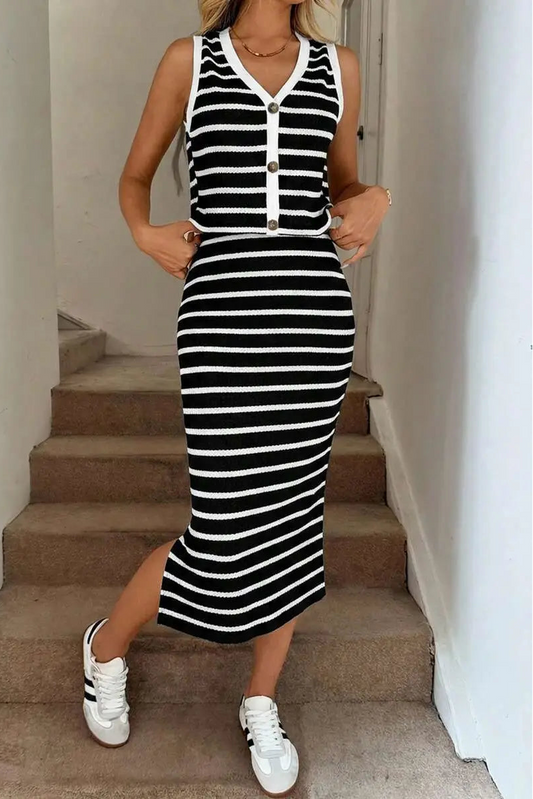 A woman wearing a sleeveless black and white horizontally striped midi dress with a V-neckline and button details, paired with white sneakers, standing on indoor concrete stairs.