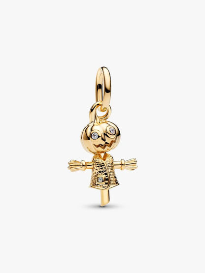 A gold charm shaped like a scarecrow, featuring a carved pumpkin head with small clear rhinestones for eyes and mouth, straw-colored arms, and a textured gold dress, shown on a light gray background.