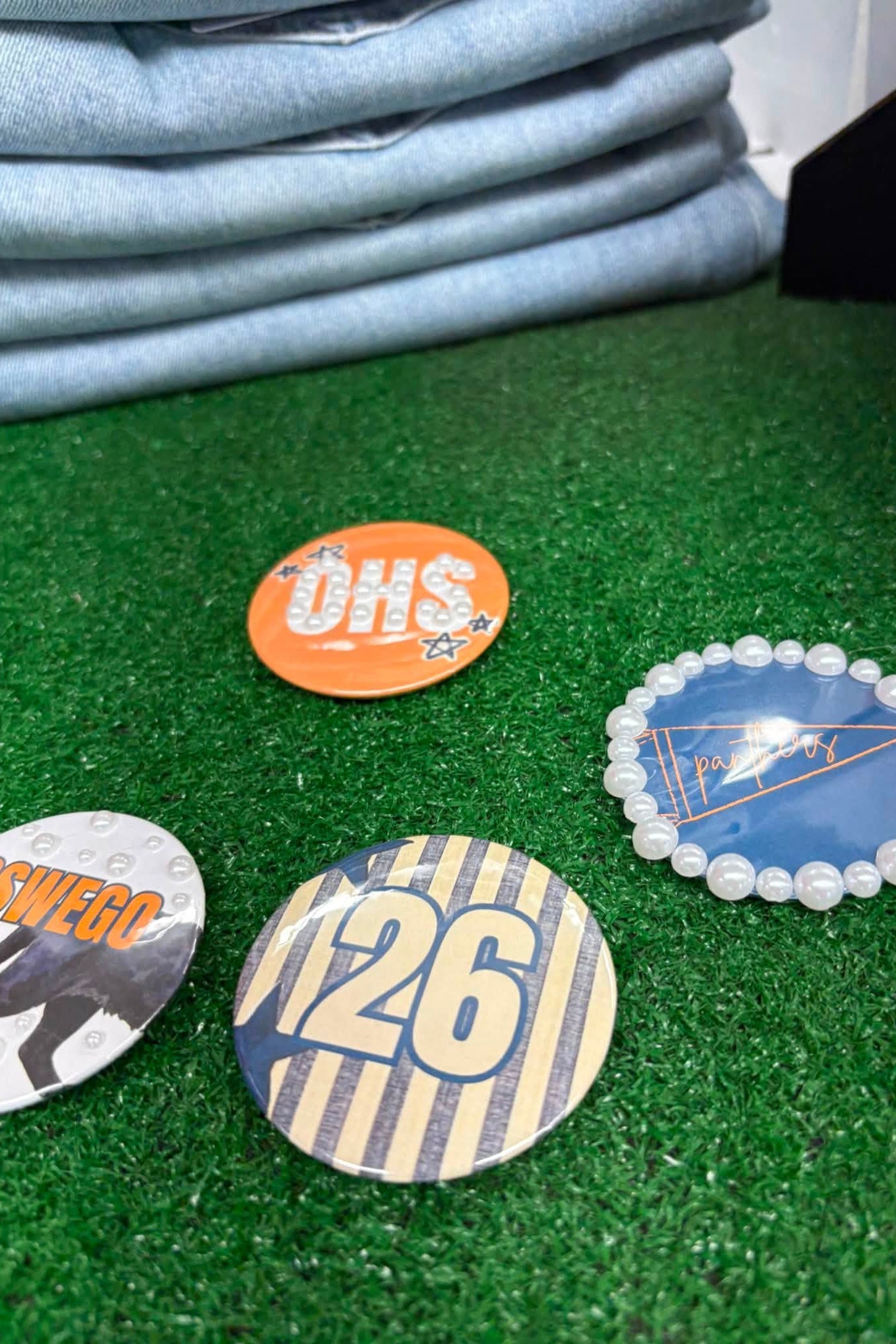 Four circular pin-back buttons displayed on green artificial turf, with a stack of light blue jeans in the background. The buttons feature various designs: one orange circle with 'DHS' in white, one blue oval with a geometric design and white pearl border, one striped beige and navy button with '26' in yellow, and a partially visible white button with orange text.